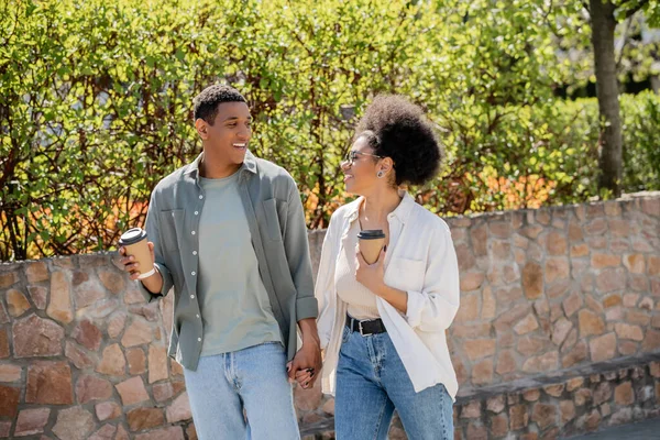 Smiling african american couple holding coffee to go while talking and walking on urban street — Stock Photo