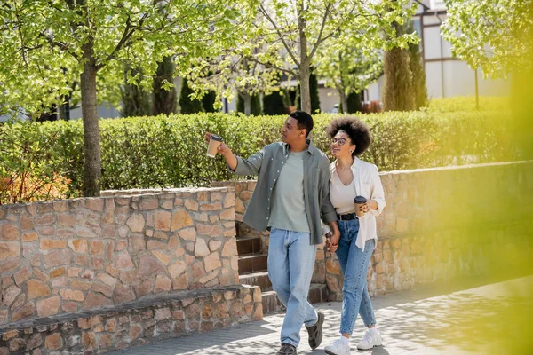 African american man with coffee to go pointing on urban street near cheerful girlfriend — Stock Photo