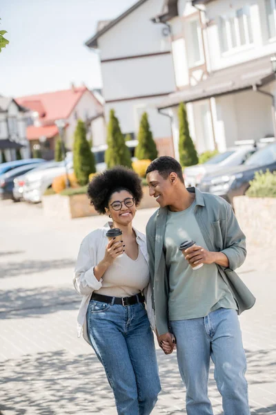 Positiv afrikanisch-amerikanisches Paar mit Kaffee zum Mitnehmen Händchen haltend beim Gehen auf der städtischen Straße — Stockfoto