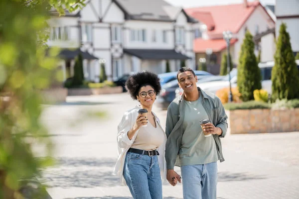 Femme afro-américaine souriante avec café pour aller marcher avec petit ami près des maisons dans la rue urbaine — Photo de stock