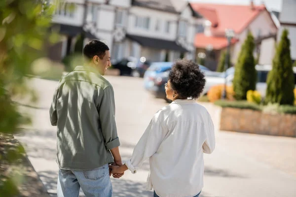Side view of cheerful african american couple talking and holding hands and walking on urban street — Stock Photo