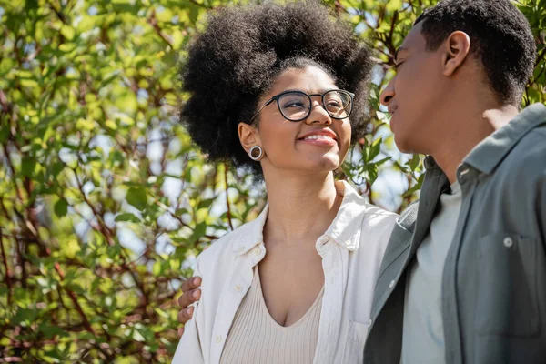 Smiling african american woman in eyeglasses looking at boyfriend on urban street in summer — Stock Photo