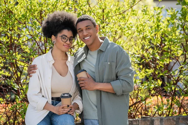 Smiling african american couple holding takeaway coffee and hugging outdoors in summer — Stock Photo