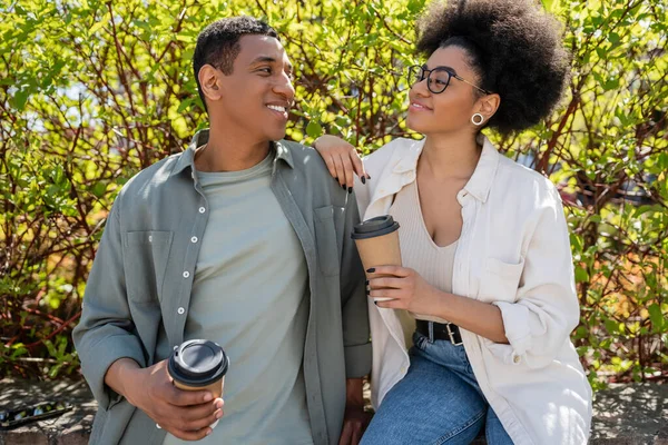 Souriant afro-américaine dans des lunettes de vue tenant du café pour aller près de petit ami à l'extérieur en été — Photo de stock