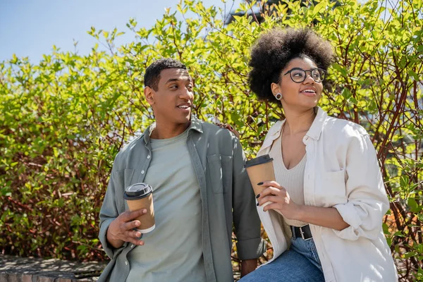 Sourire couple afro-américain tenant du café pour aller près des plantes et des arbres à l'extérieur en été — Photo de stock