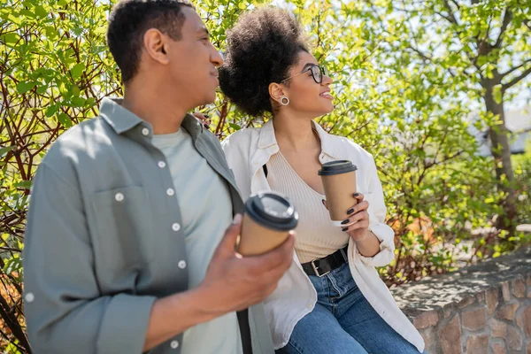 Усміхнена афроамериканка тримає каву, щоб піти ближче розмитий хлопець і дерева на відкритому повітрі — стокове фото