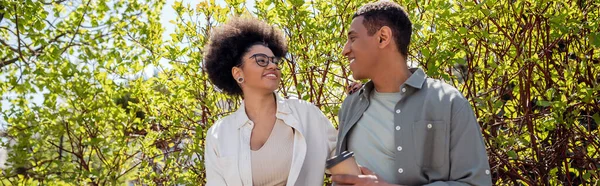 Uomo afroamericano sorridente con caffè per andare a parlare con la ragazza vicino agli alberi all'aperto, striscione — Foto stock
