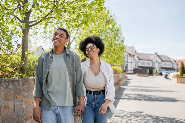 Positive african american couple holding hands while walking on urban street in summer — Stock Photo