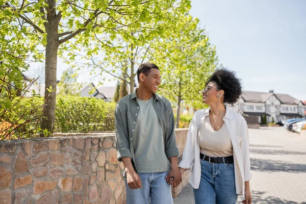 Sonriente pareja afroamericana cogida de la mano y hablando mientras camina por la calle urbana en verano - foto de stock