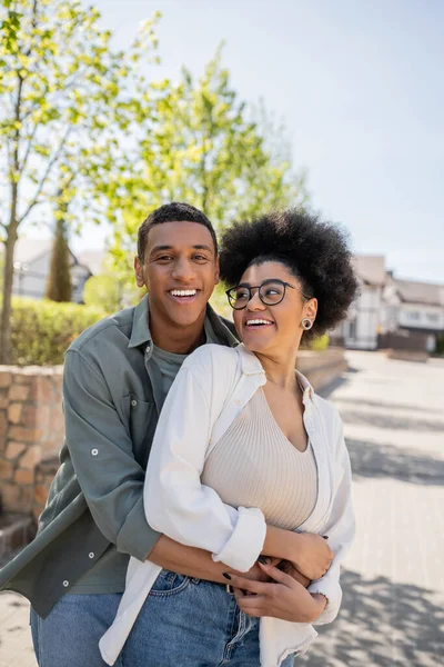 Sorridente afro-americano homem abraçando namorada em óculos e olhando para a câmera na rua urbana — Fotografia de Stock