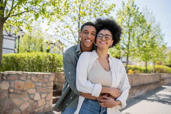 African american man embracing girlfriend in eyeglasses while standing together outdoors in summer — Stock Photo