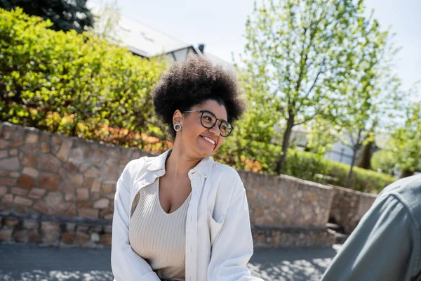Smiling african american woman in eyeglasses looking at blurred boyfriend on street in summer — Stock Photo