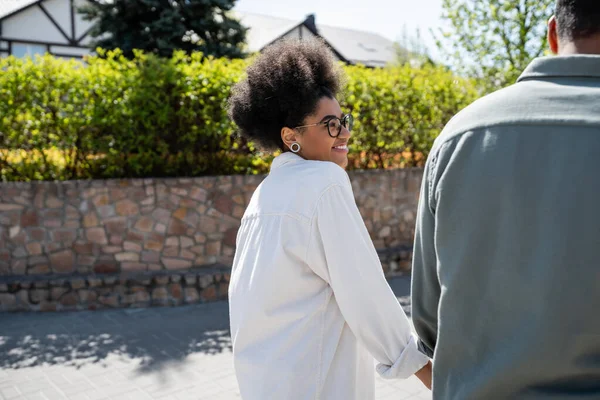 Улыбающаяся африканская американка в рубашке и очках, смотрящая на парня на городской улице — стоковое фото