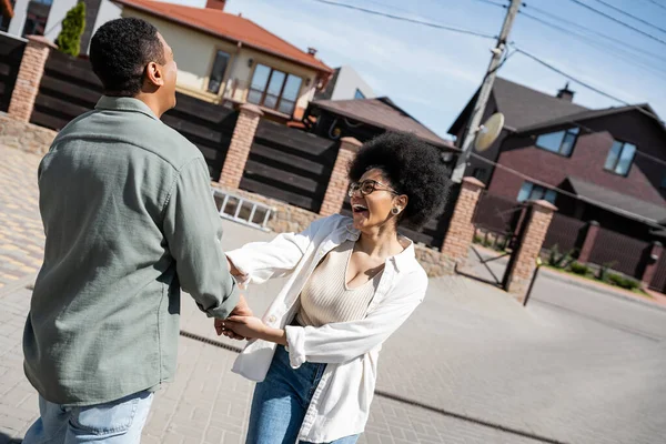 Happy african american couple holding hands while standing near blurred houses on street — Stock Photo