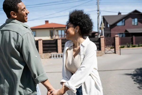 Vista lateral do casal afro-americano sorridente de mãos dadas na rua urbana com casas no fundo — Fotografia de Stock