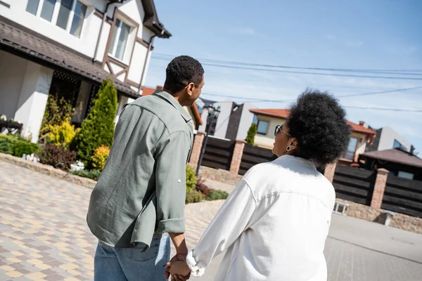 Freudig afrikanisch-amerikanisches Paar hält Händchen in der Nähe von verschwommenem Haus auf der Straße im Sommer — Stockfoto