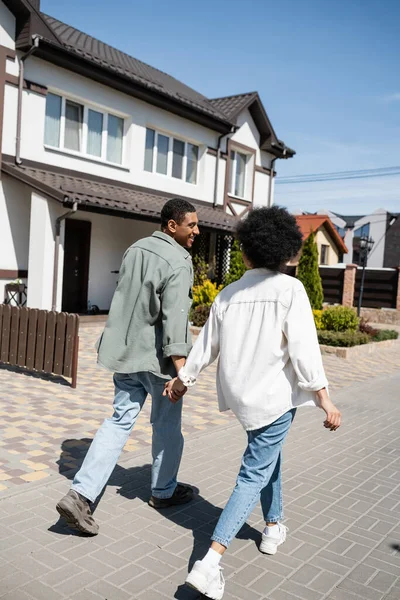Smiling african american couple holding hands while walking on sidewalk near houses on street — Stock Photo