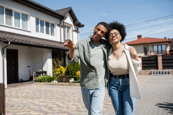 Positive african american couple hugging and walking near blurred houses on urban street — Stock Photo