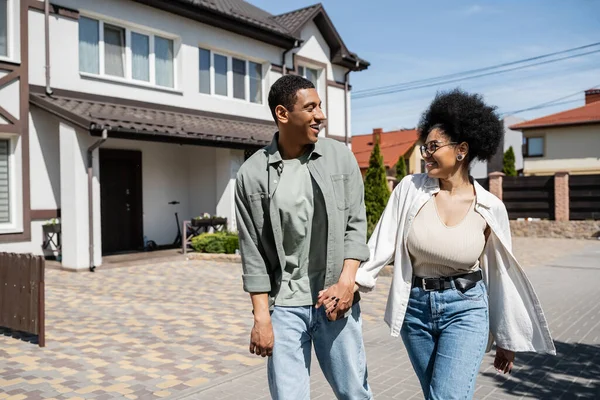 Alegre pareja afroamericana cogida de la mano mientras camina por la calle cerca de las casas en verano - foto de stock