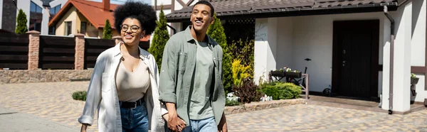 Alegre casal afro-americano de mãos dadas e caminhando juntos perto de casas na rua, bandeira — Fotografia de Stock