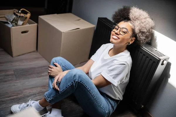 Smiling african american woman looking at camera near cardboard boxes in new house — Stock Photo