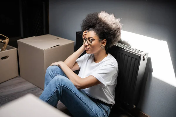 Positive afrikanisch-amerikanische Frau mit Brille sitzt neben Pappschachteln in neuem Haus — Stockfoto