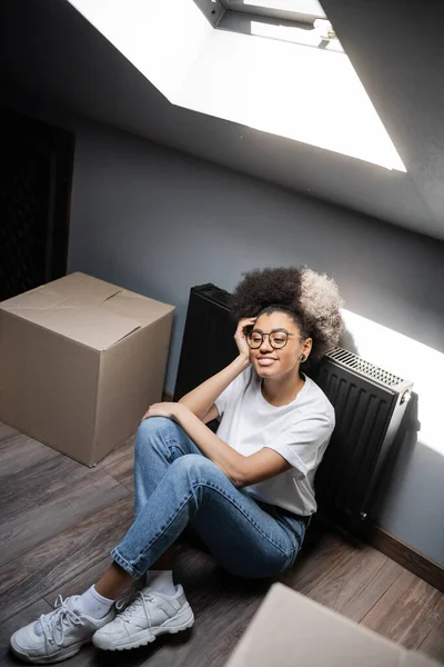 Hochwinkelaufnahme einer lächelnden afrikanisch-amerikanischen Frau, die in der Nähe von Kartons auf dem Dachboden in einem Haus sitzt — Stockfoto