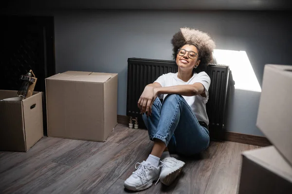 Heureuse femme afro-américaine assise au soleil près des boîtes en carton sur le grenier dans une nouvelle maison — Photo de stock