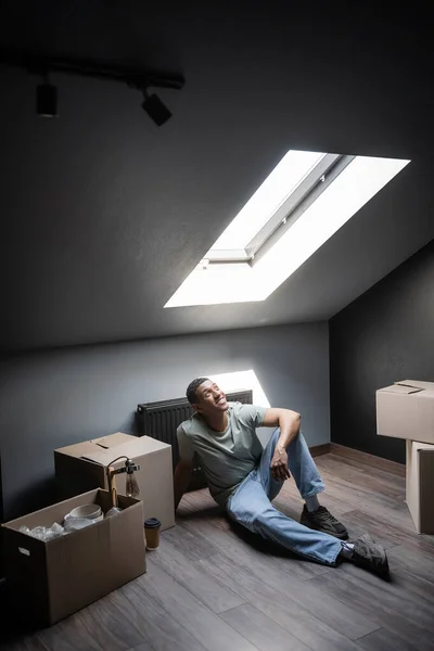 Fröhlicher afrikanisch-amerikanischer Mann sitzt im Sonnenlicht in der Nähe von Kartons auf dem Dachboden in neuem Haus — Stockfoto