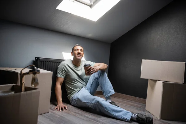 Sorrindo afro-americano homem segurando café para ir perto de caixas de papelão no sótão em casa nova — Fotografia de Stock