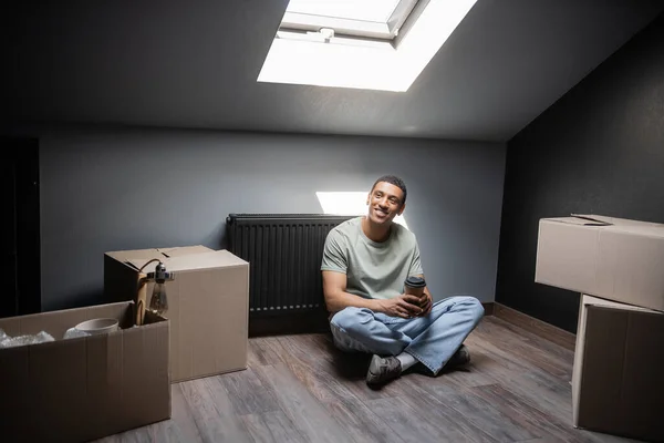 Joyful african american man holding coffee to go near carton boxes on attic in new home — Stock Photo