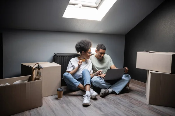Sonriente africano americano pareja usando portátil y tarjeta de crédito cerca de cajas en ático en nueva casa - foto de stock