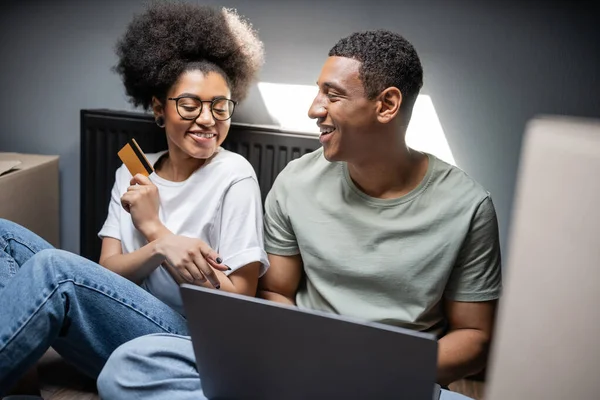 Весела афроамериканська пара використовує ноутбук і кредитну картку біля картонних коробок в новому будинку — стокове фото