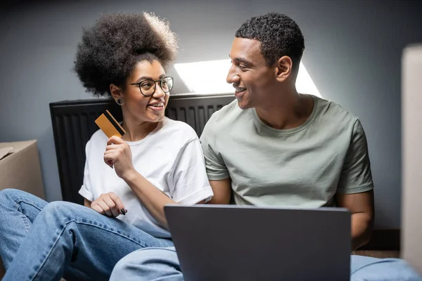 Усміхнена афроамериканка тримає кредитну картку біля хлопця з ноутбуком у новому будинку — стокове фото