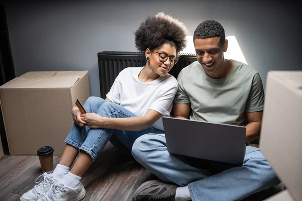 Pareja afroamericana positiva usando el ordenador portátil y la tarjeta de crédito en el ático en la casa nueva - foto de stock