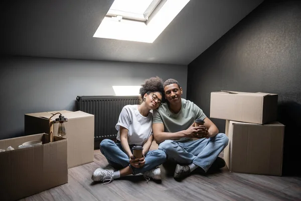 Усміхнена афро-американська пара тримає каву, щоб піти, сидячи біля коробок на горищі в новому будинку — стокове фото