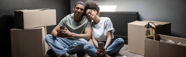 Alegre casal afro-americano segurando takeaway café perto de caixas de papelão na nova casa, banner — Fotografia de Stock