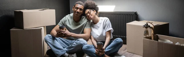 Alegre pareja afroamericana celebración de café para llevar cerca de cajas de cartón en casa nueva, pancarta - foto de stock