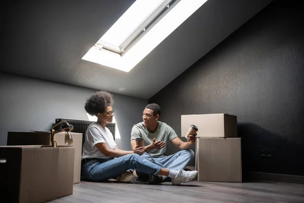 Uomo afroamericano sorridente con caffè per andare a parlare con la ragazza vicino scatole di cartone in una nuova casa — Foto stock