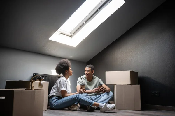 Радісна афроамериканська пара з кавою піде розмовляти біля картонних коробок на горищі в новому будинку — стокове фото