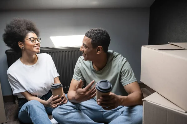 Hombre afroamericano positivo hablando con su novia con café para ir al ático en casa nueva - foto de stock