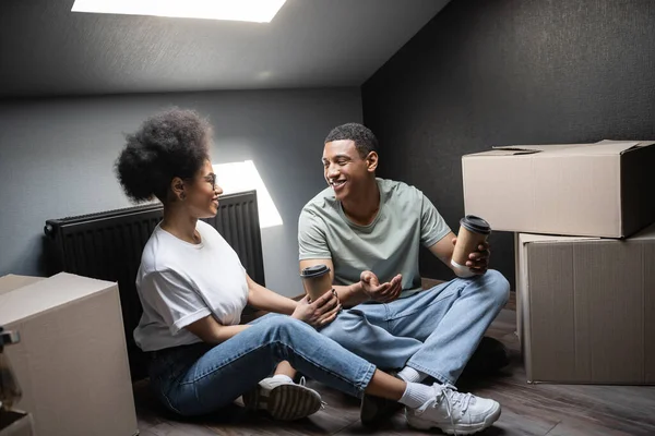 Alegre casal afro-americano com café falando perto de caixas de papelão no sótão na nova casa — Fotografia de Stock