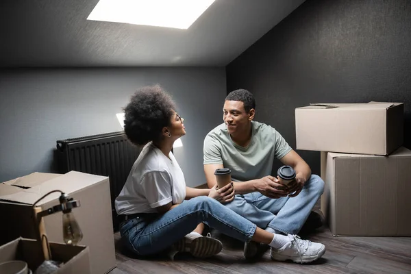 African american woman with coffee to go talking to boyfriend near boxes on attic in new house — Stock Photo