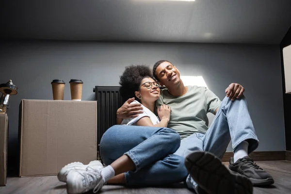 Alegre casal afro-americano abraçando perto de café na caixa de papelão no sótão na nova casa — Fotografia de Stock
