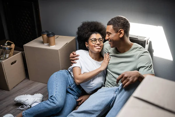 Cheerful african american woman hugging boyfriend near coffee to go and boxes in new house — Stock Photo