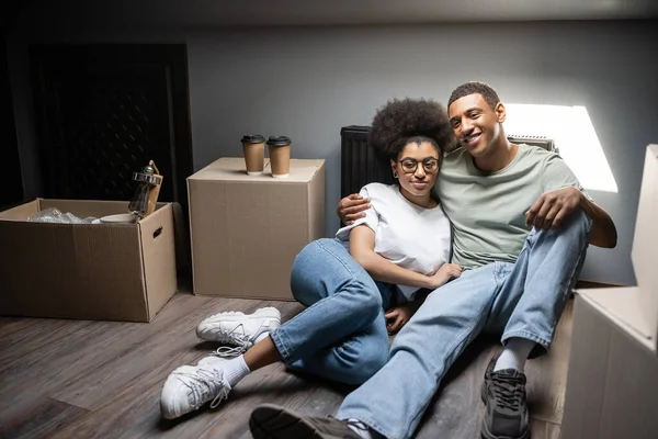 Улыбающаяся романтическая африканская пара, обнимающаяся возле кофе и коробки на чердаке в новом доме — стоковое фото