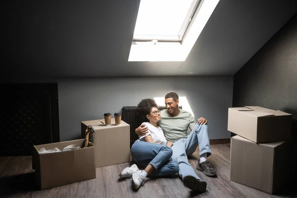 Positive african american couple hugging near cardboard boxes and coffee to go in new house — Stock Photo