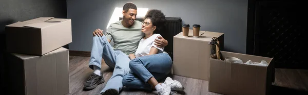 Усміхнена афро-американська пара обіймається біля картонних коробок і виймає каву в новому будинку, банер — стокове фото