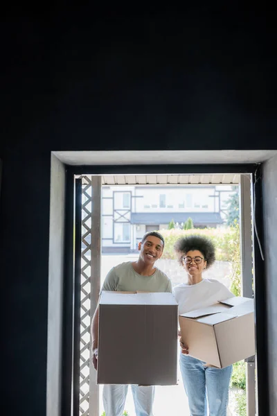 Sorrindo casal afro-americano segurando caixas de papelão e olhando para a câmera perto da porta na nova casa — Fotografia de Stock