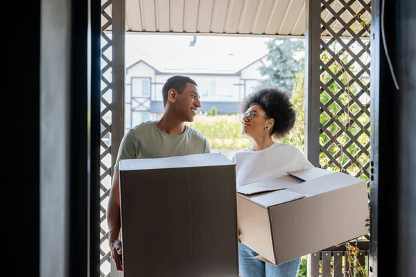 Sorridente romântico casal afro-americano segurando caixas de papelão perto da porta da nova casa — Fotografia de Stock
